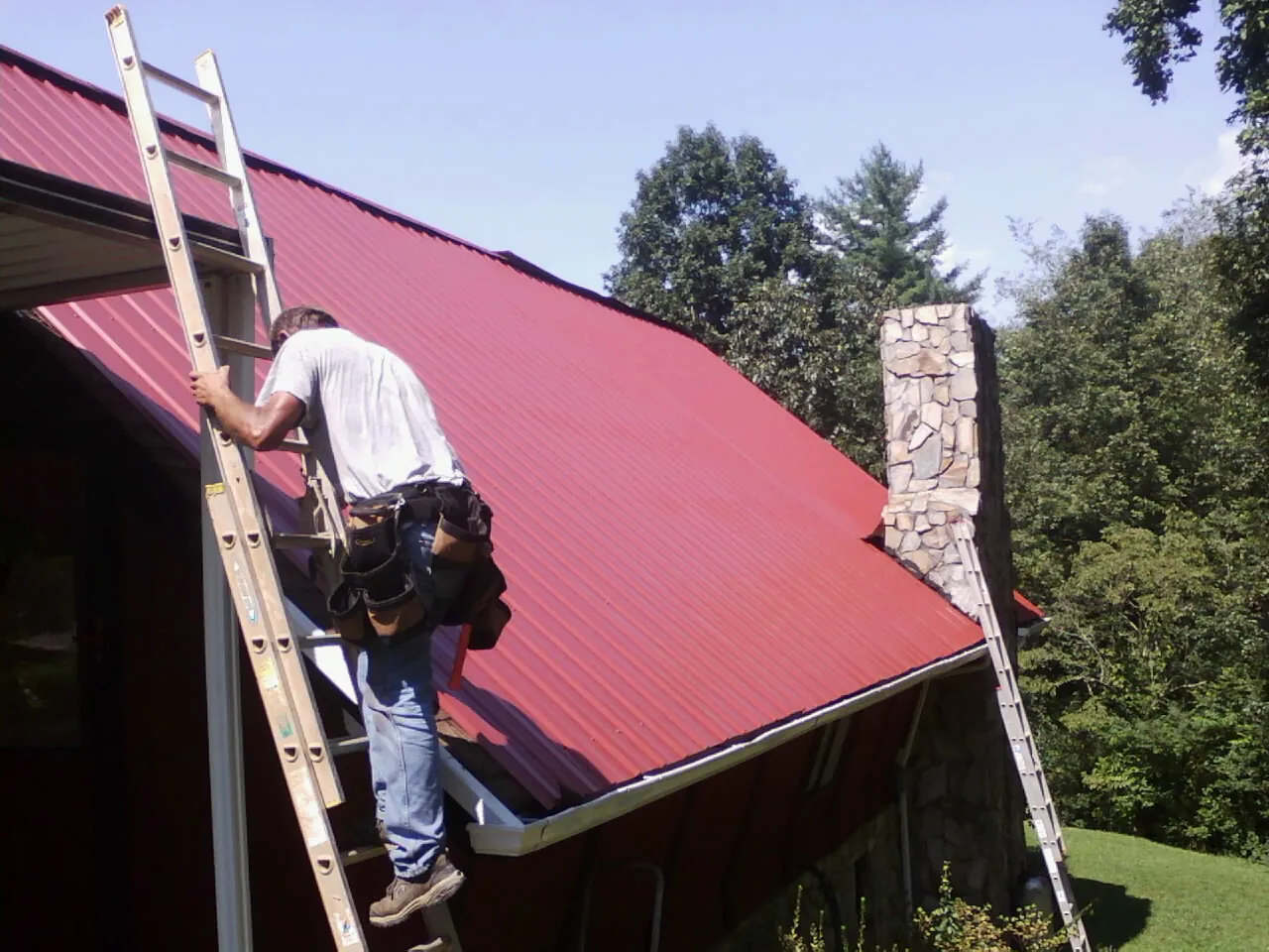 Roofer on a ladder beside a red metal roof and stone chimney — Soffit & Fascia Repair by Roofing Brothers in Palenville